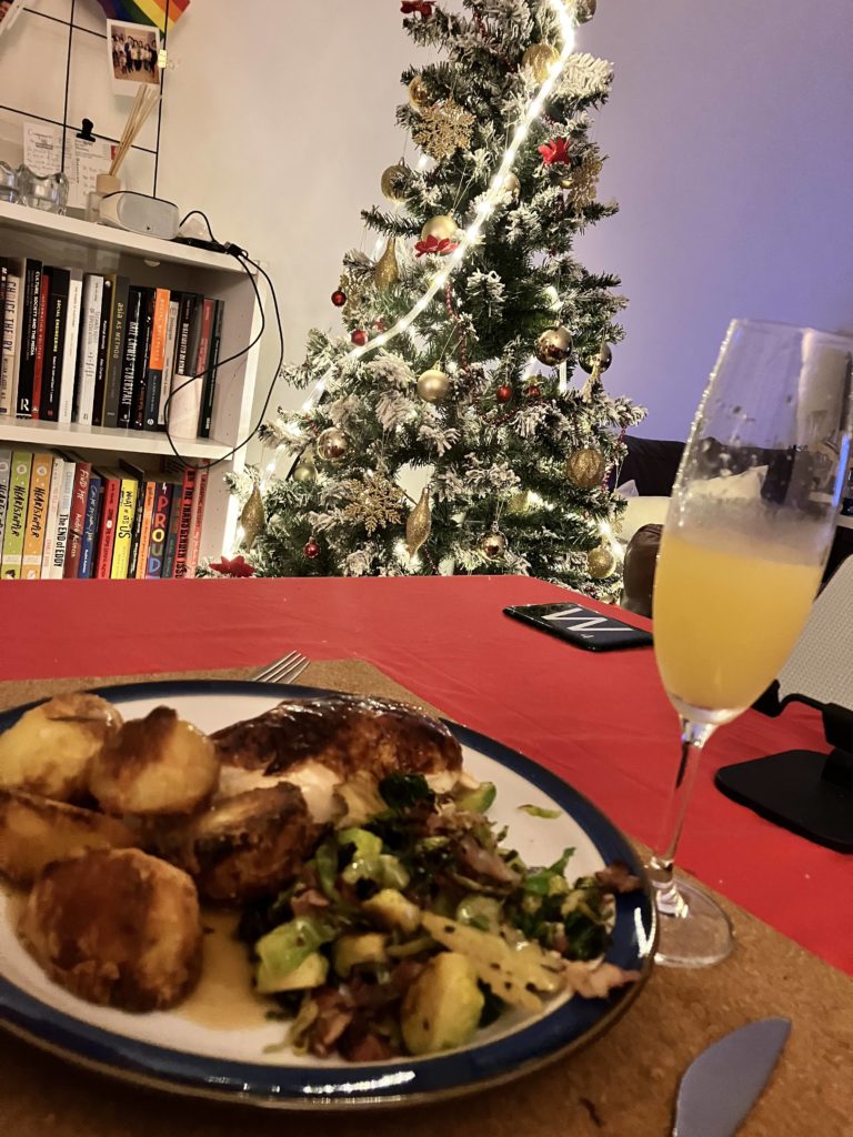 A roast dinner on a plate next to a glass of Buck's Fizz, with a bookshelf and Christmas tree in the background.