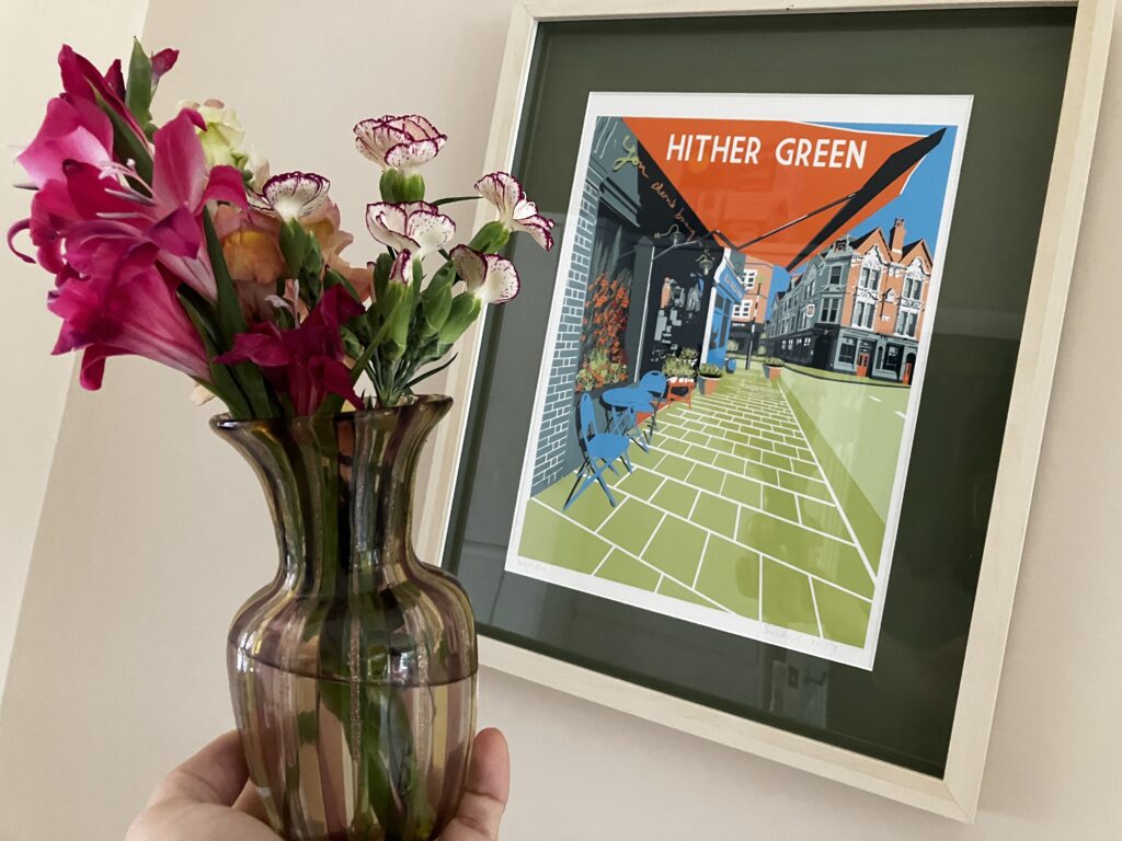 A hand holding up a vase of flowers next to an artwork featuring a street in Hither Green in London.